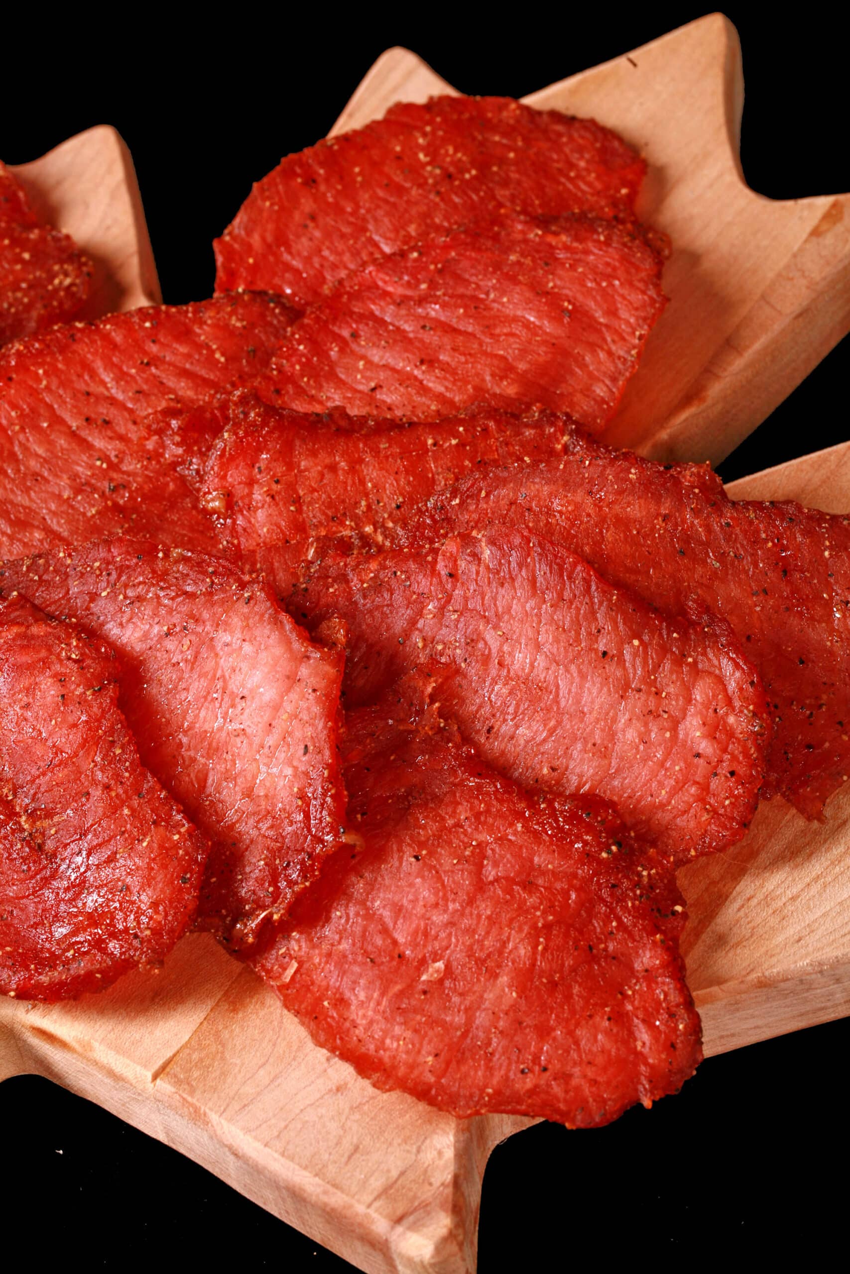 A pile of back bacon jerky slices on a maple leaf shaped wooden cutting board.