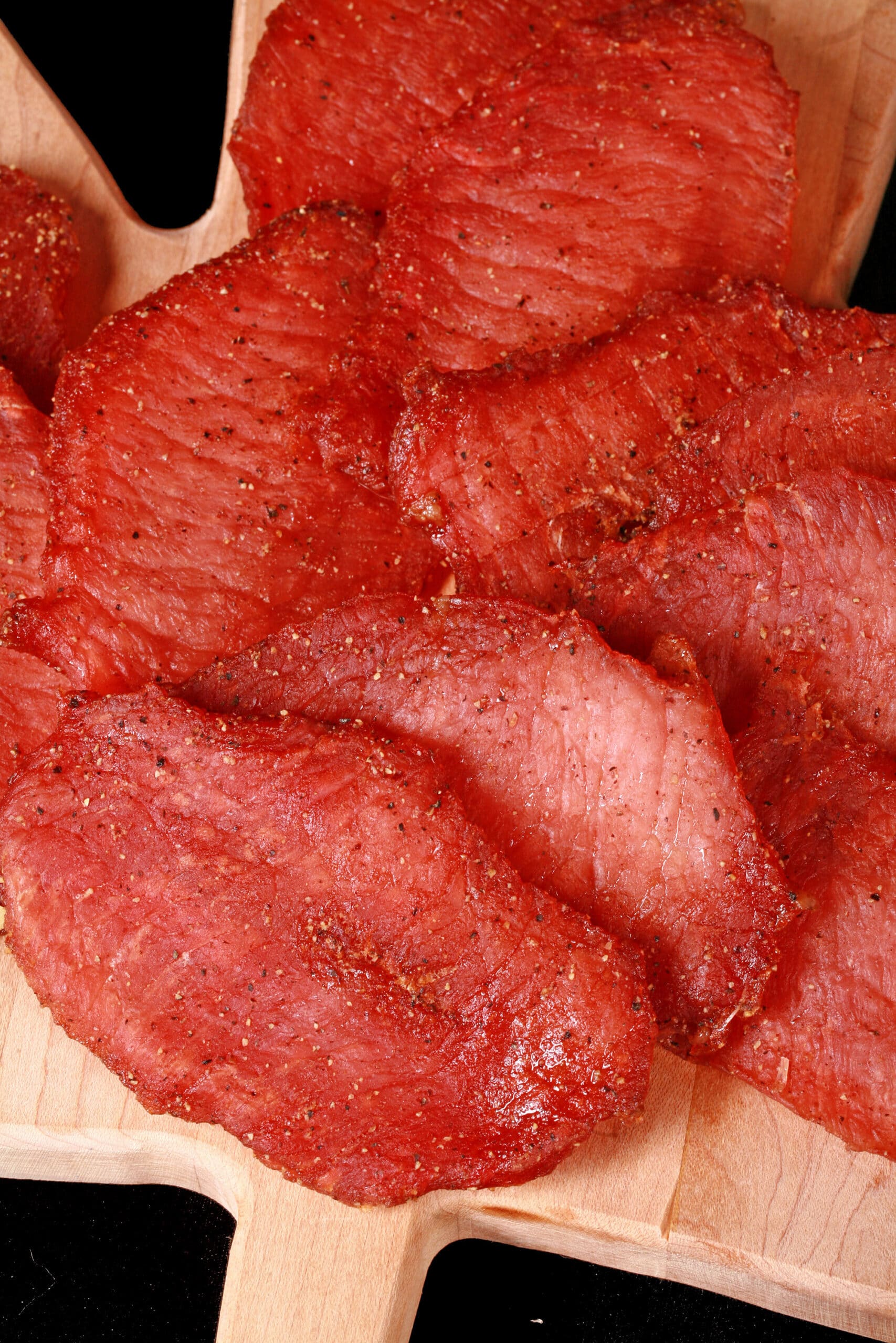 A pile of back bacon jerky slices on a maple leaf shaped wooden cutting board.
