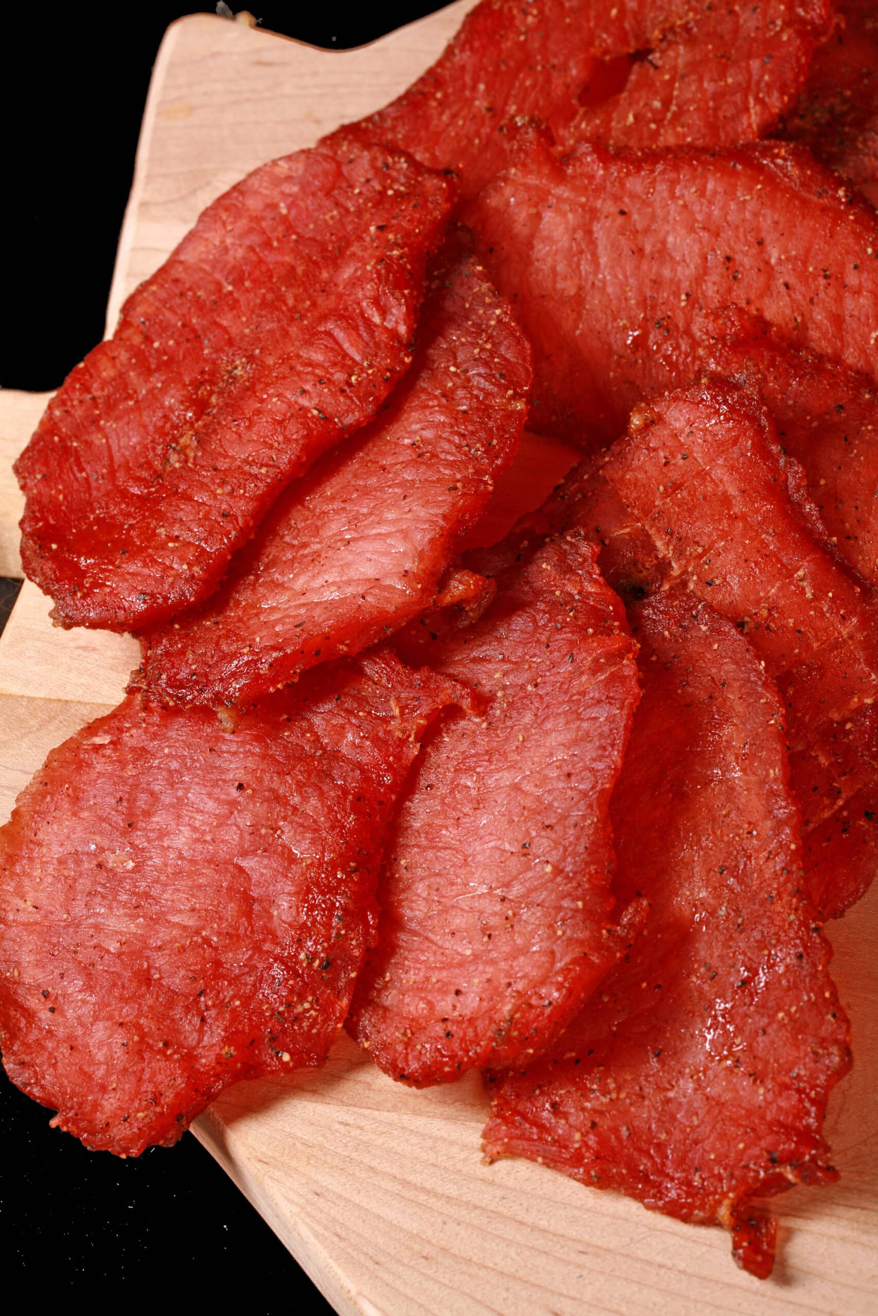 A pile of back bacon jerky slices on a maple leaf shaped wooden cutting board.
