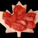 A pile of back bacon jerky slices on a maple leaf shaped wooden cutting board.