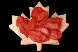 A pile of back bacon jerky slices on a maple leaf shaped wooden cutting board.