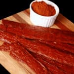 Several strips of berbere chicken jerky on a cutting board with a ramekin of berbere seasoning.