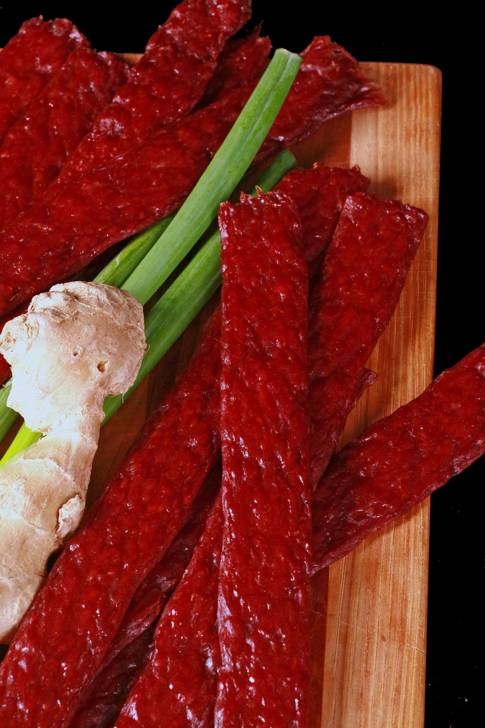Several strips of teriyaki beef jerky on a cutting board with a knob of ginger and some green onions.