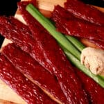 Several strips of teriyaki beef jerky on a cutting board with a knob of ginger and some green onions.