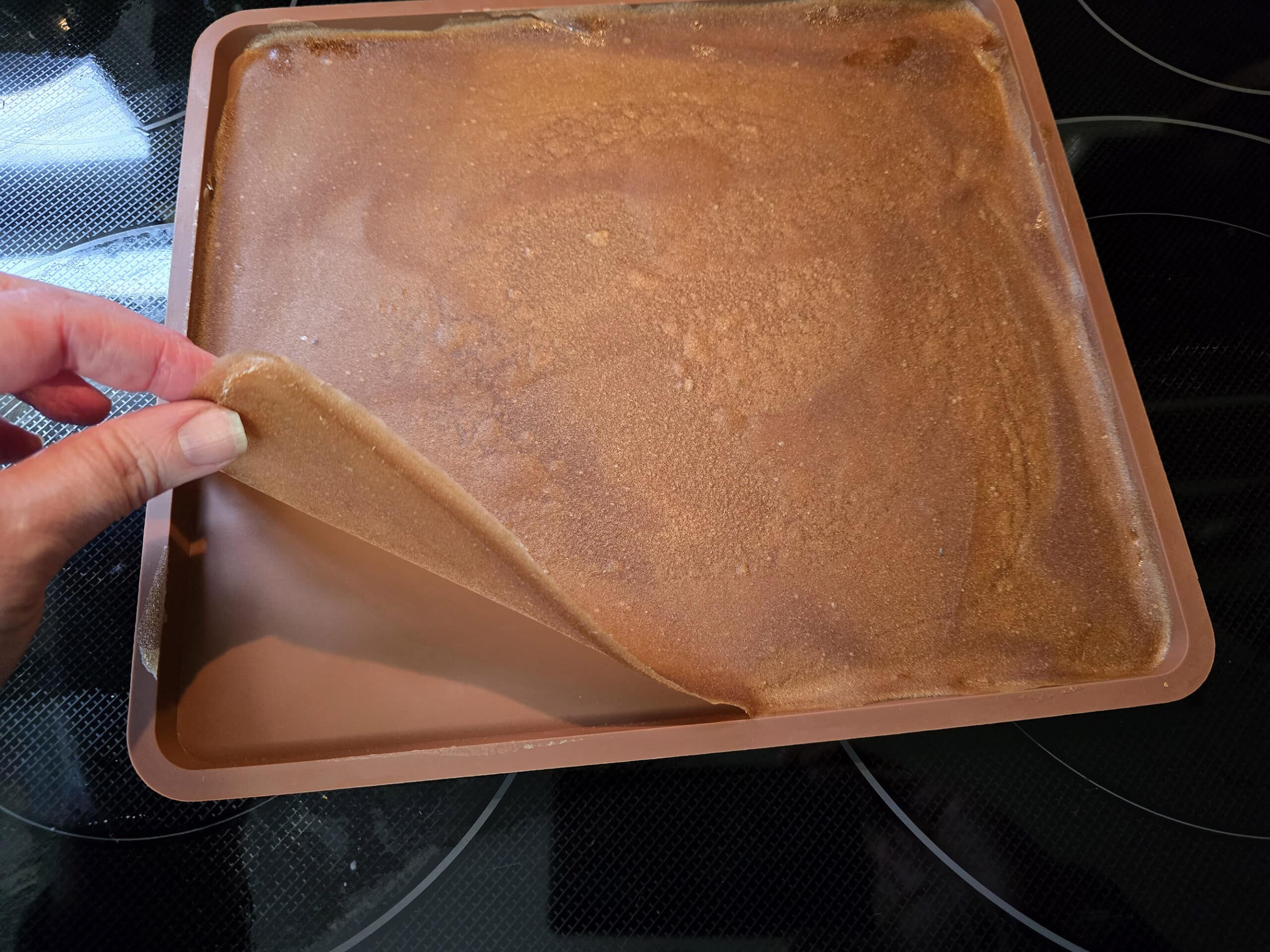 A hand pulls up a corner of the apple cinnamon fruit leather, away from the silicone tray.