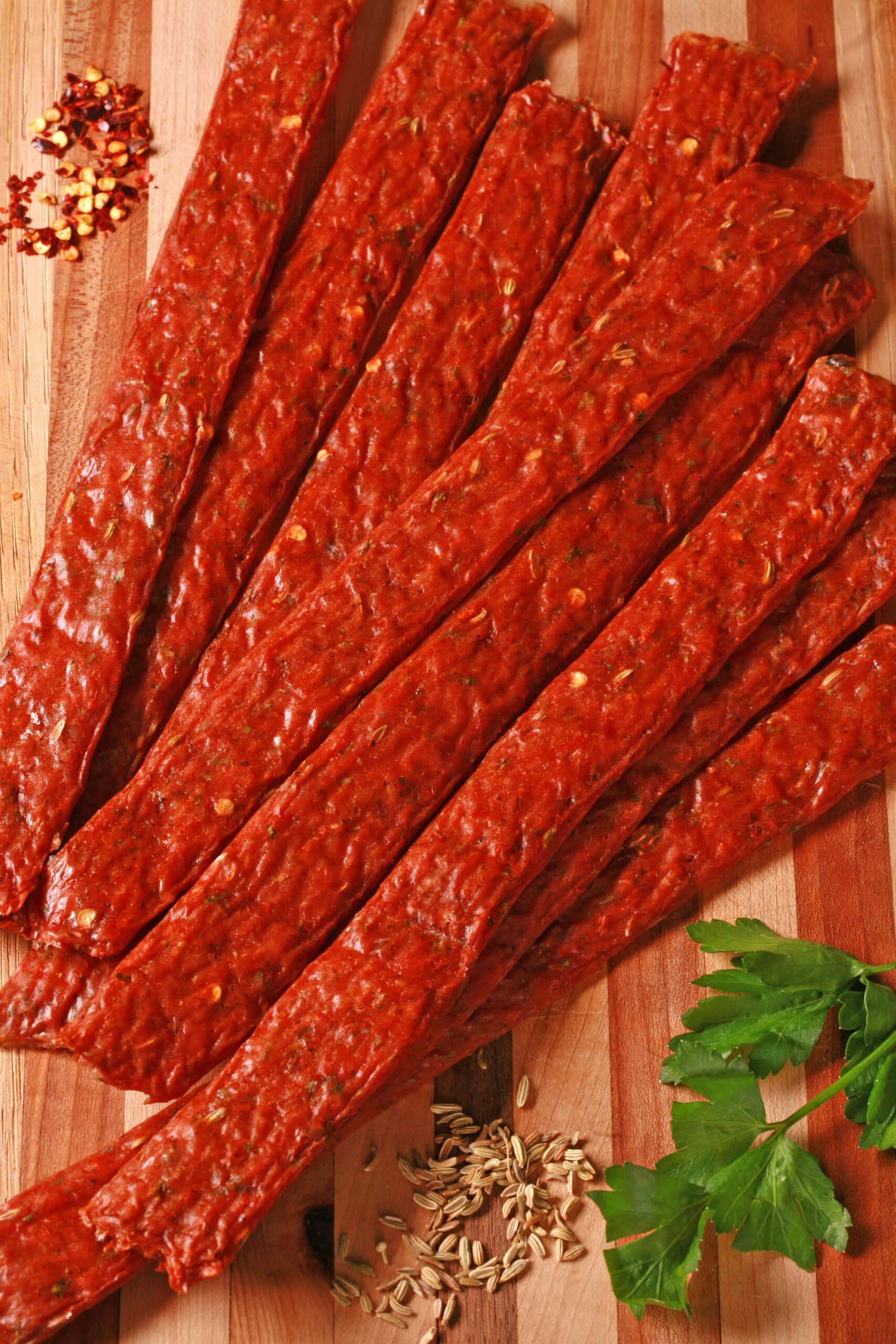 Several strips of Italian Sausage ground meat jerky on a cutting board with fresh parsley and fennel seeds.