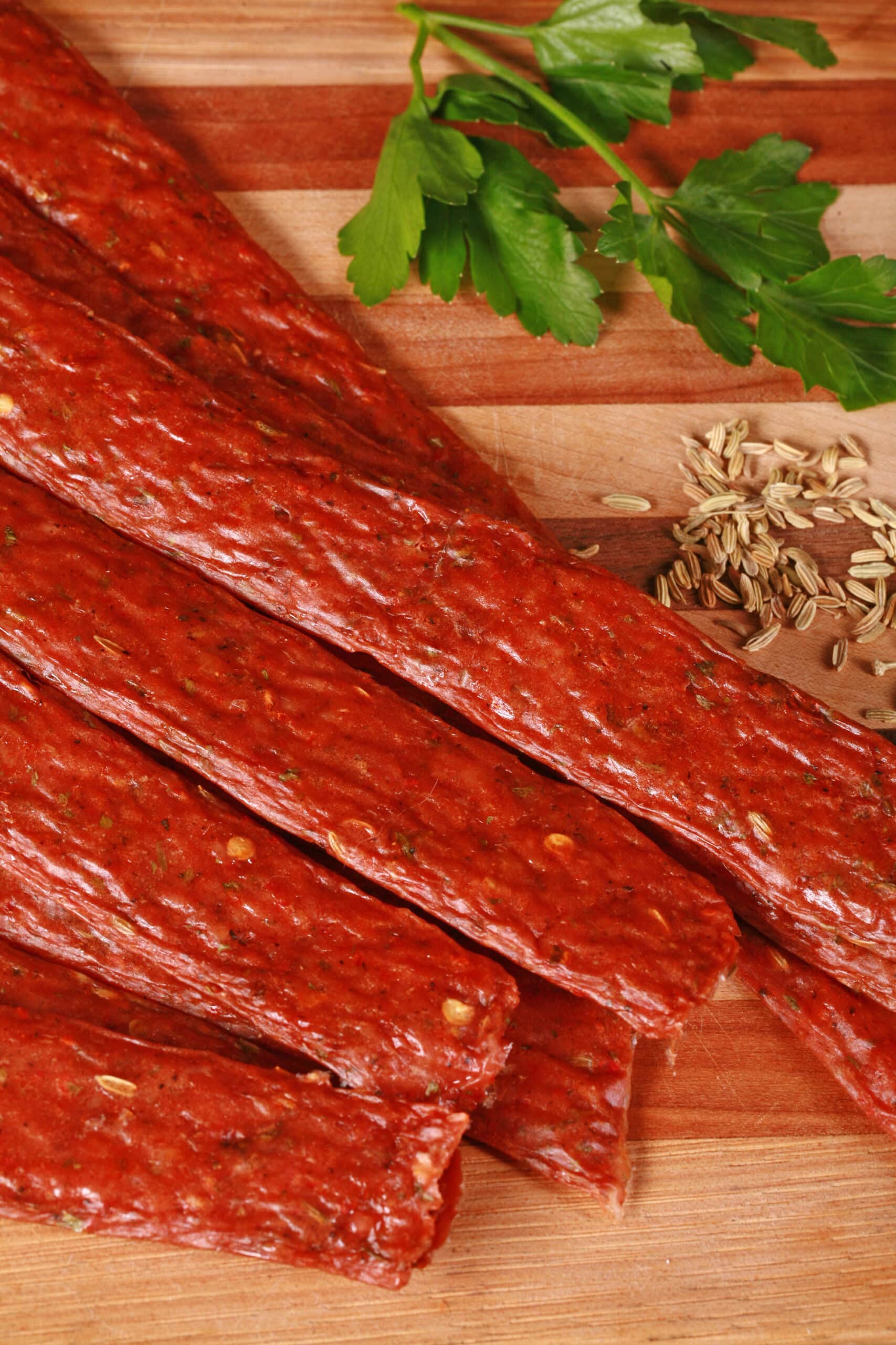Several strips of Italian Sausage ground meat jerky on a cutting board with fresh parsley and fennel seeds.