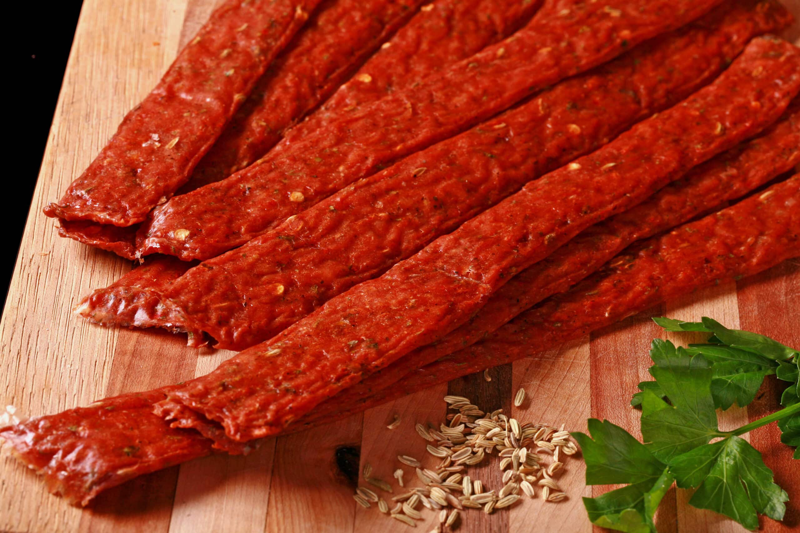 Several strips of Italian Sausage ground meat jerky on a cutting board with fresh parsley and fennel seeds.