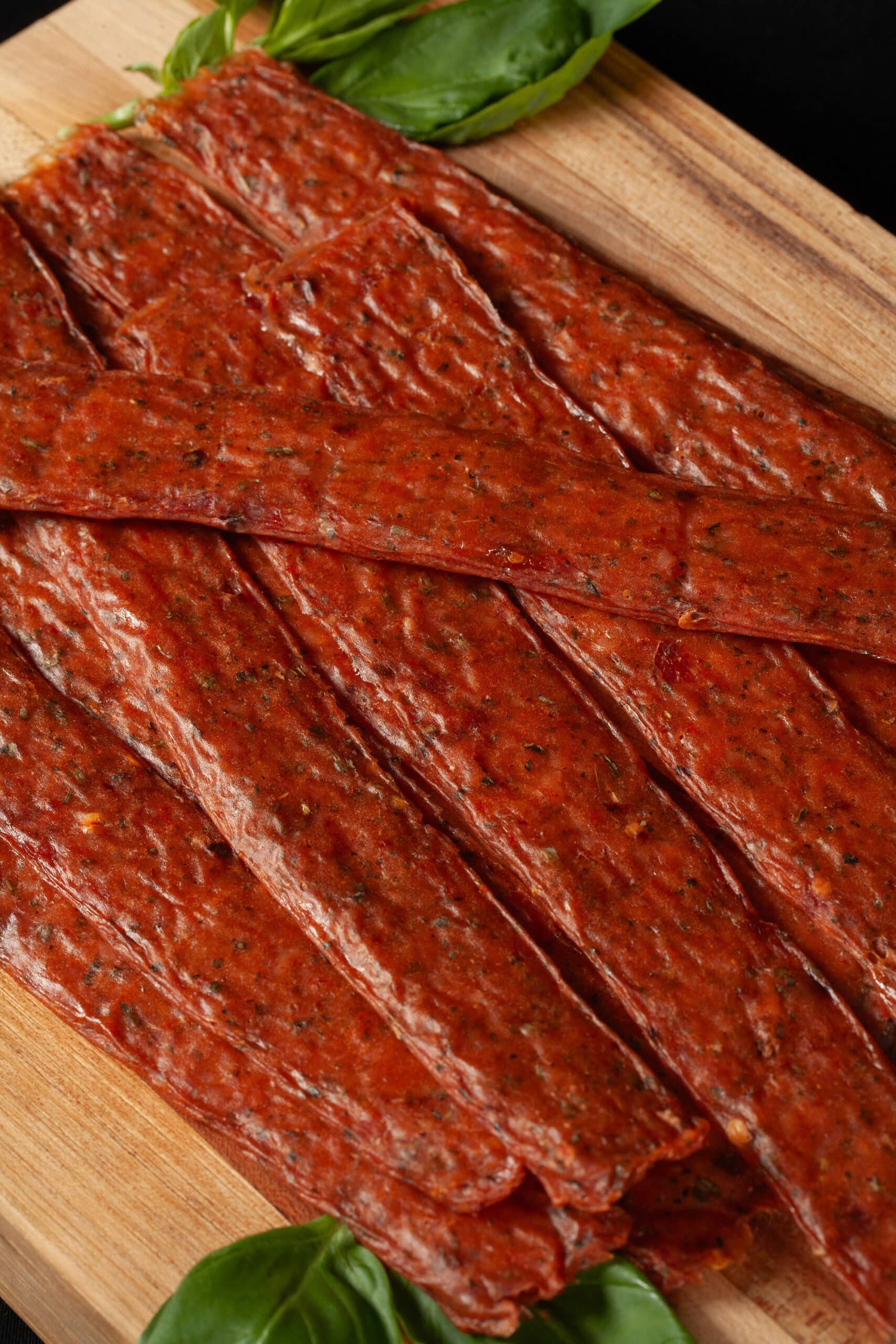 A pile of pizza jerky strips on a cutting board, with some fresh basil.