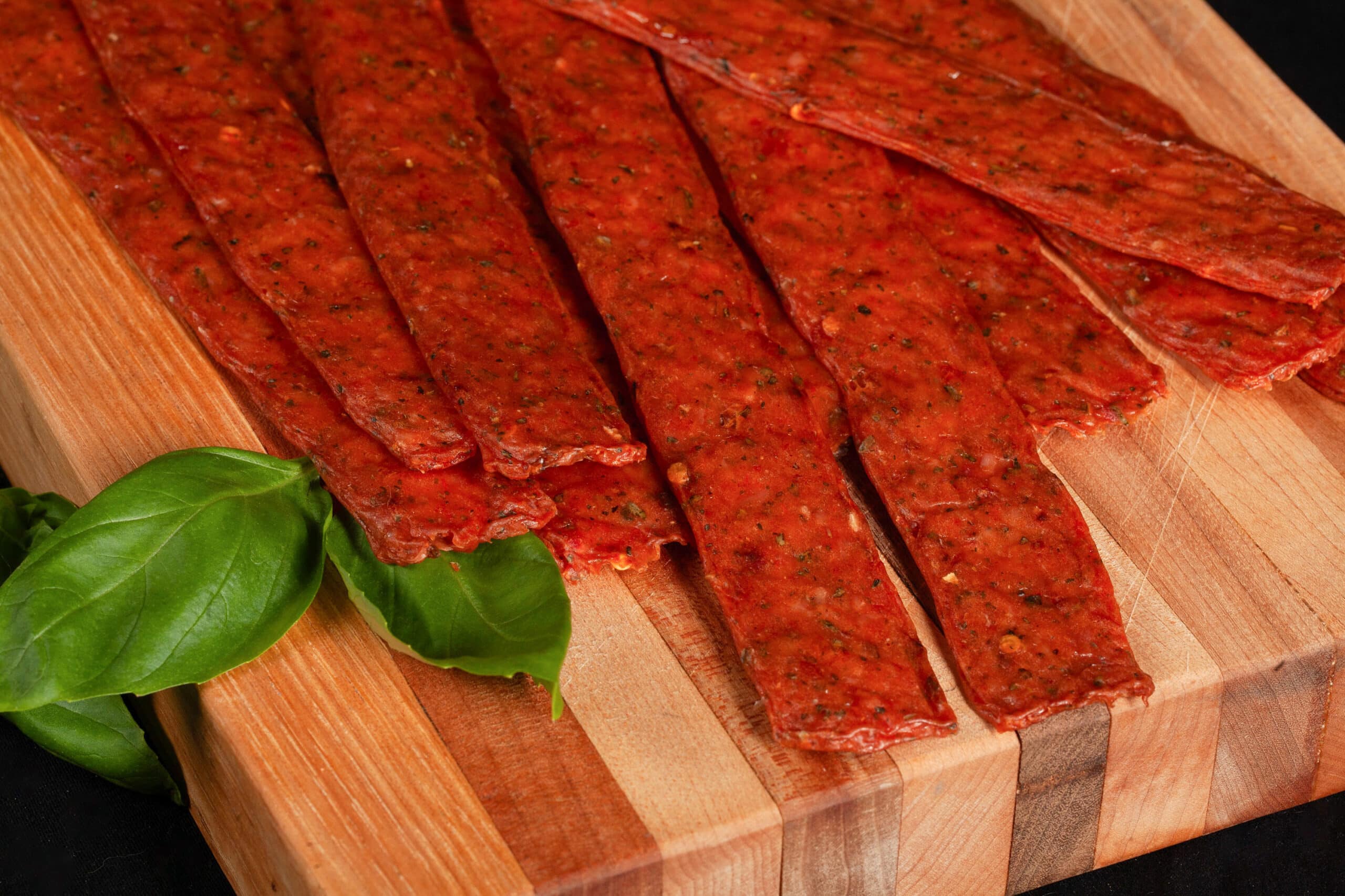 A pile of pizza jerky strips on a cutting board, with some fresh basil.