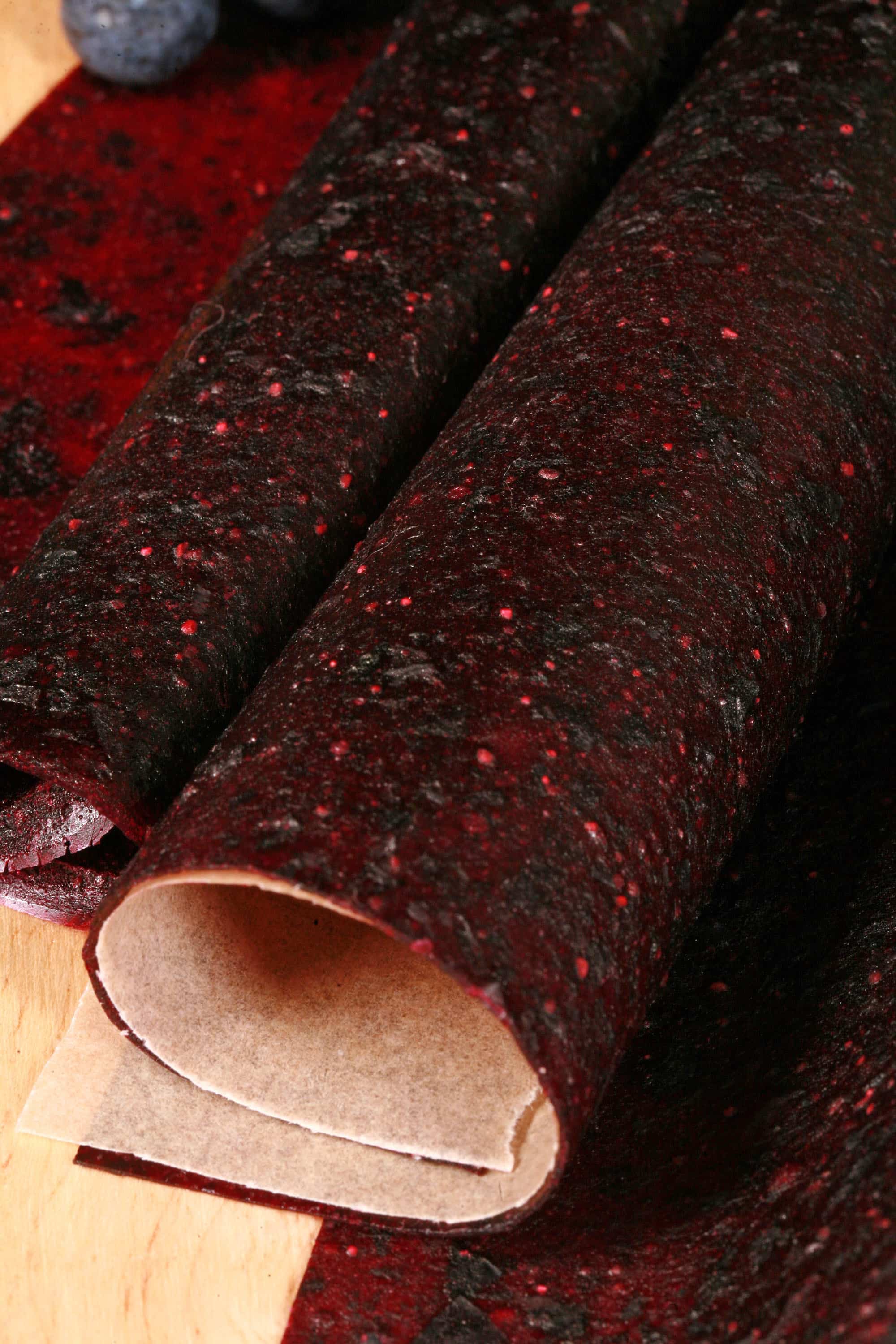 Several homemade Apple Blueberry fruit leather rolls on a cutting board, with a green apple and some fresh blueberries.