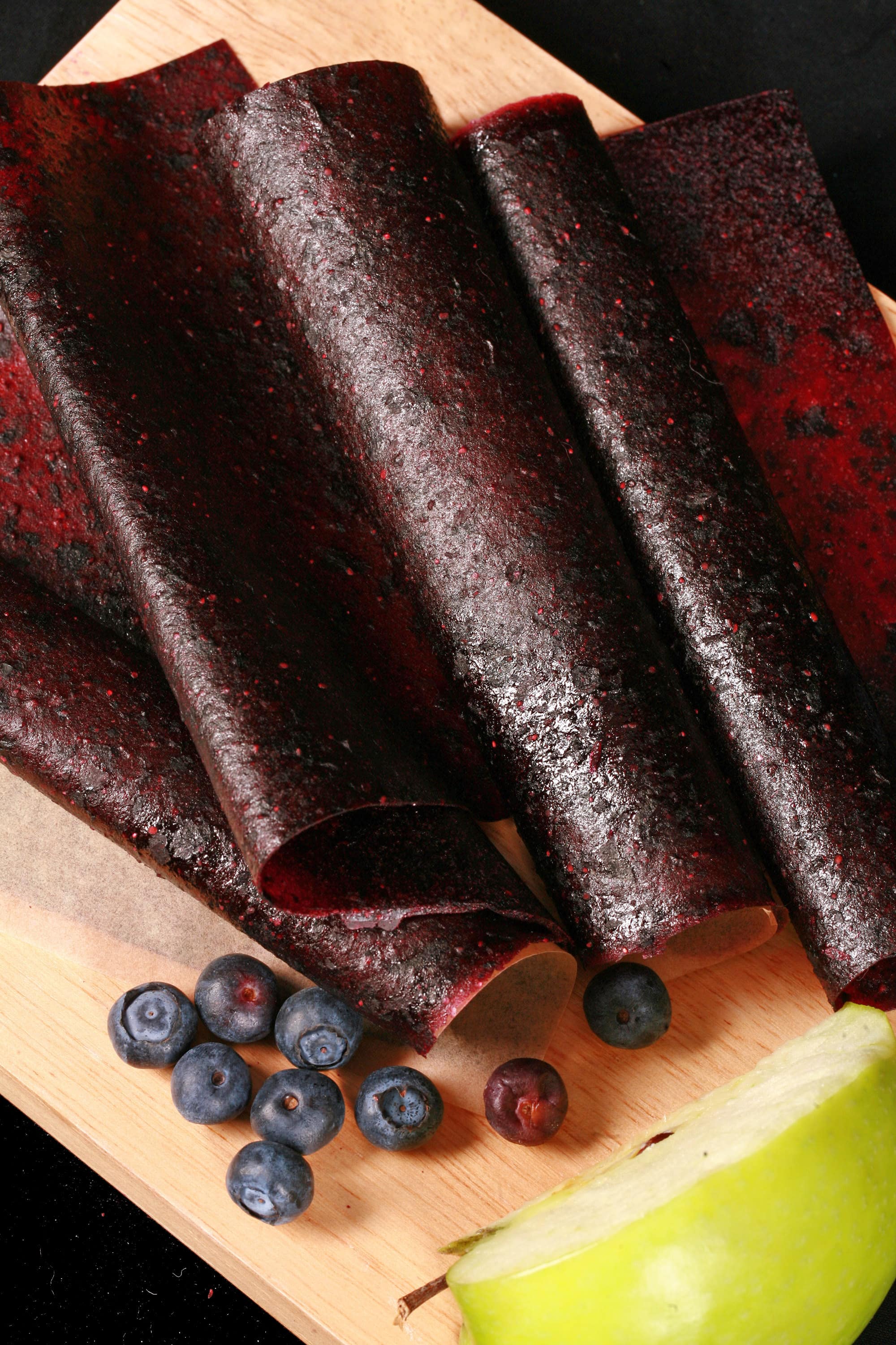 Several homemade blueberry apple fruit leather rolls on a cutting board, with a green apple and some fresh blueberries.