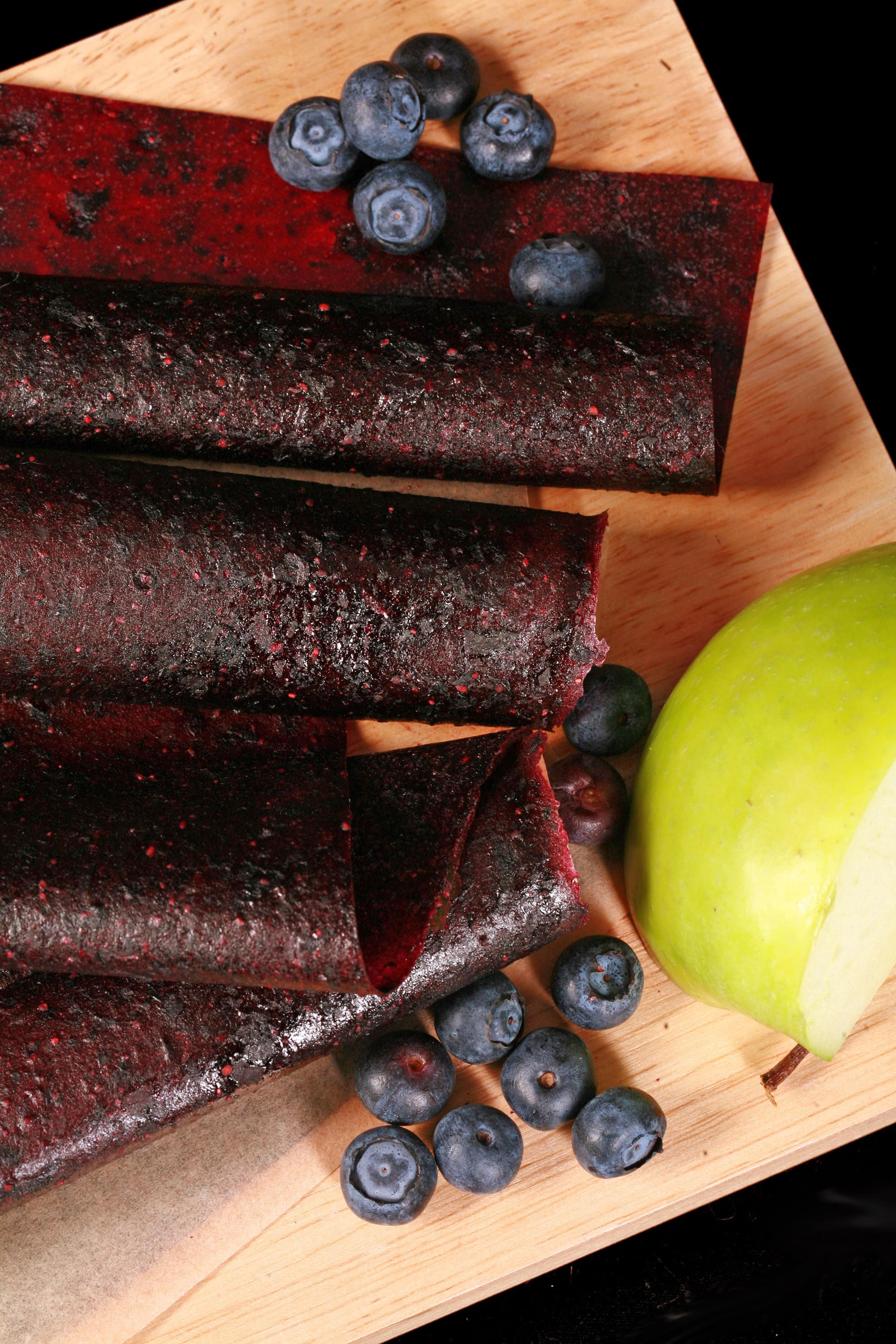 Several homemade Apple Blueberry fruit leather rolls on a cutting board, with a green apple and some fresh blueberries.