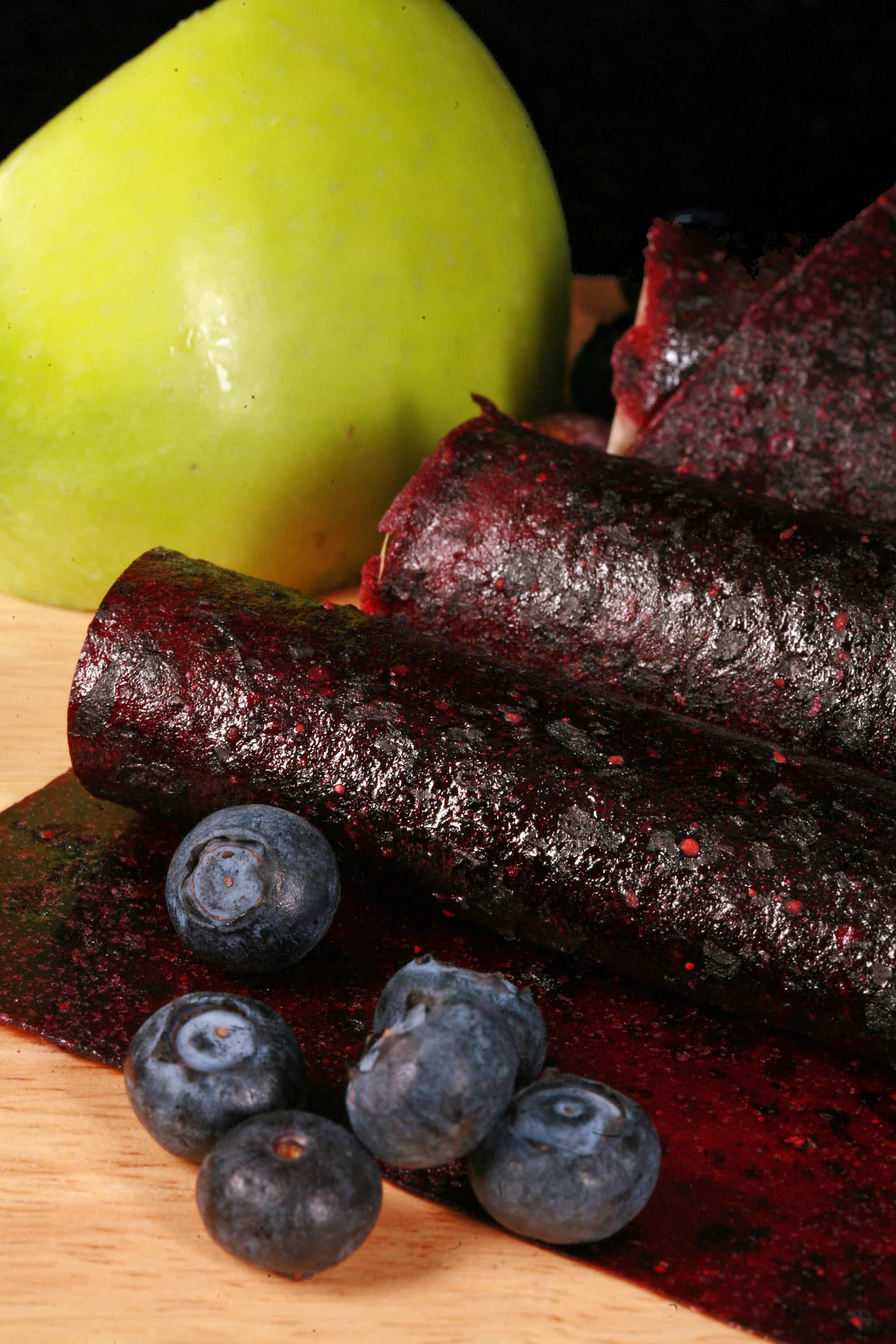 Several homemade blueberry apple fruit leather rolls on a cutting board, with a green apple and some fresh blueberries.