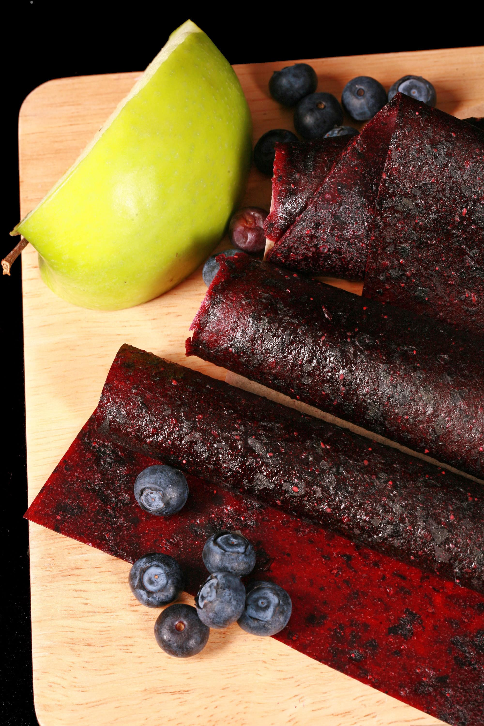 Several homemade Apple Blueberry fruit leather rolls on a cutting board, with a green apple and some fresh blueberries.
