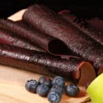 Several homemade blueberry apple fruit leather rolls on a cutting board, with a green apple and some fresh blueberries.