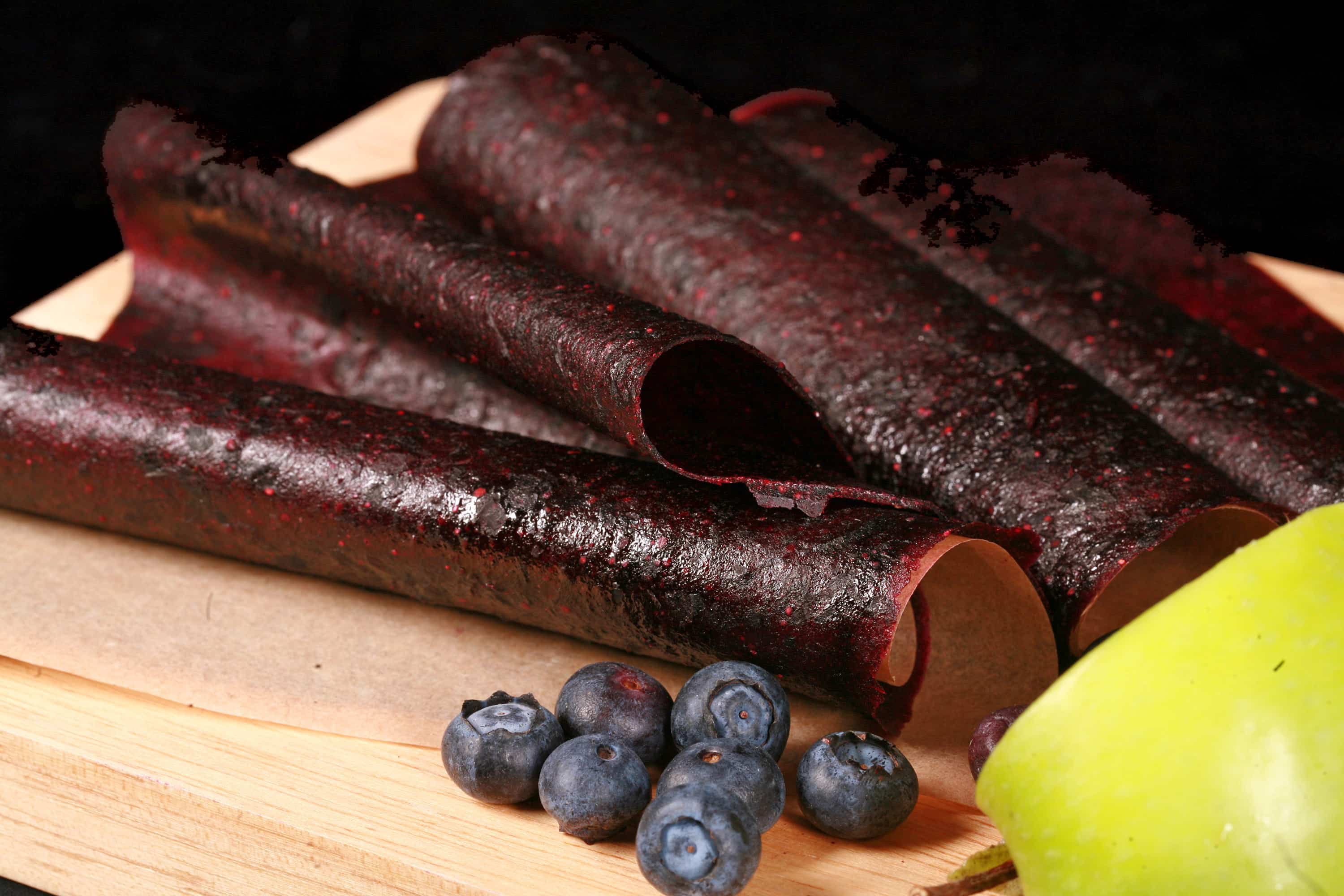 Several homemade blueberry apple fruit leather rolls on a cutting board, with a green apple and some fresh blueberries.