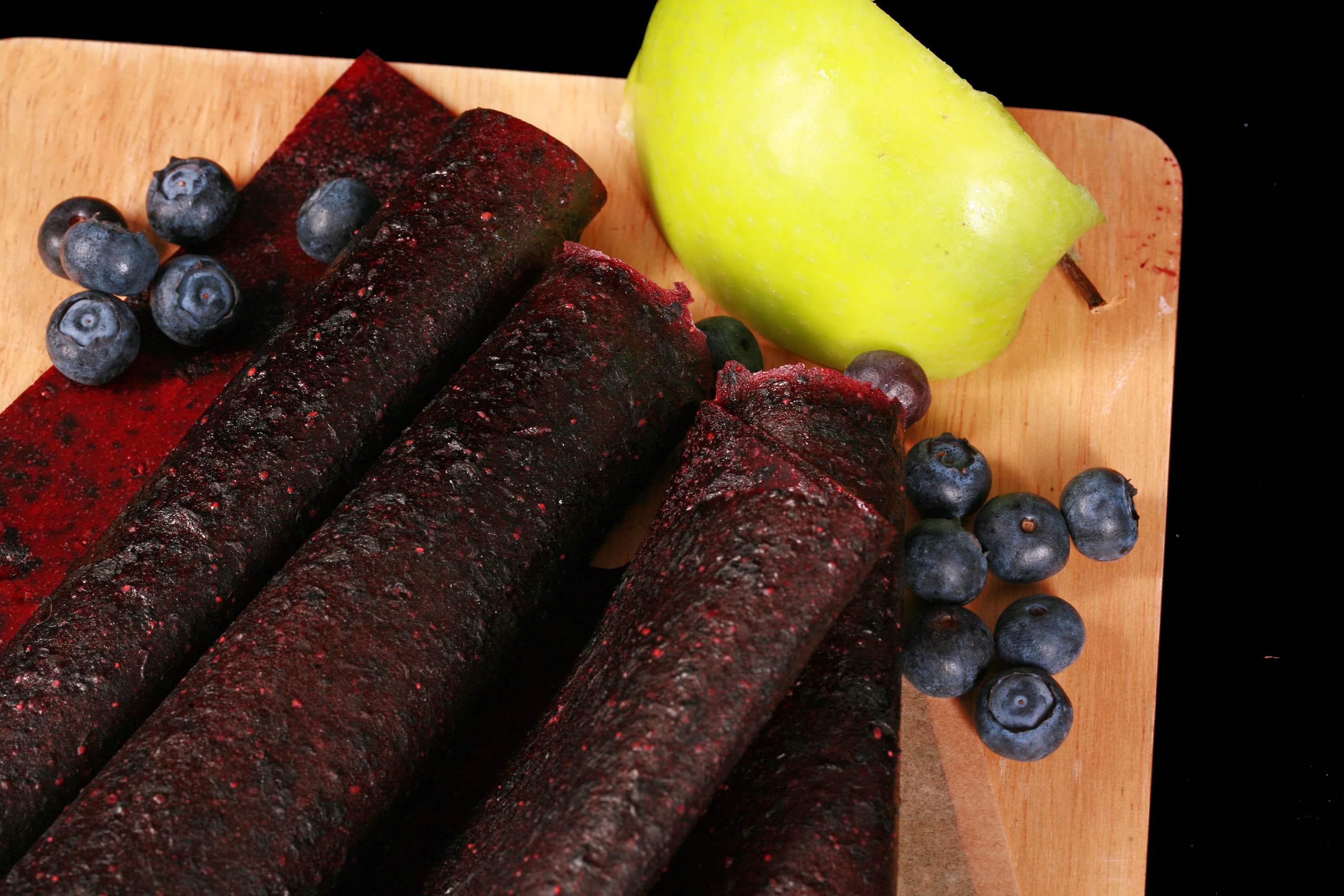 Several homemade Apple Blueberry fruit leather rolls on a cutting board, with a green apple and some fresh blueberries.