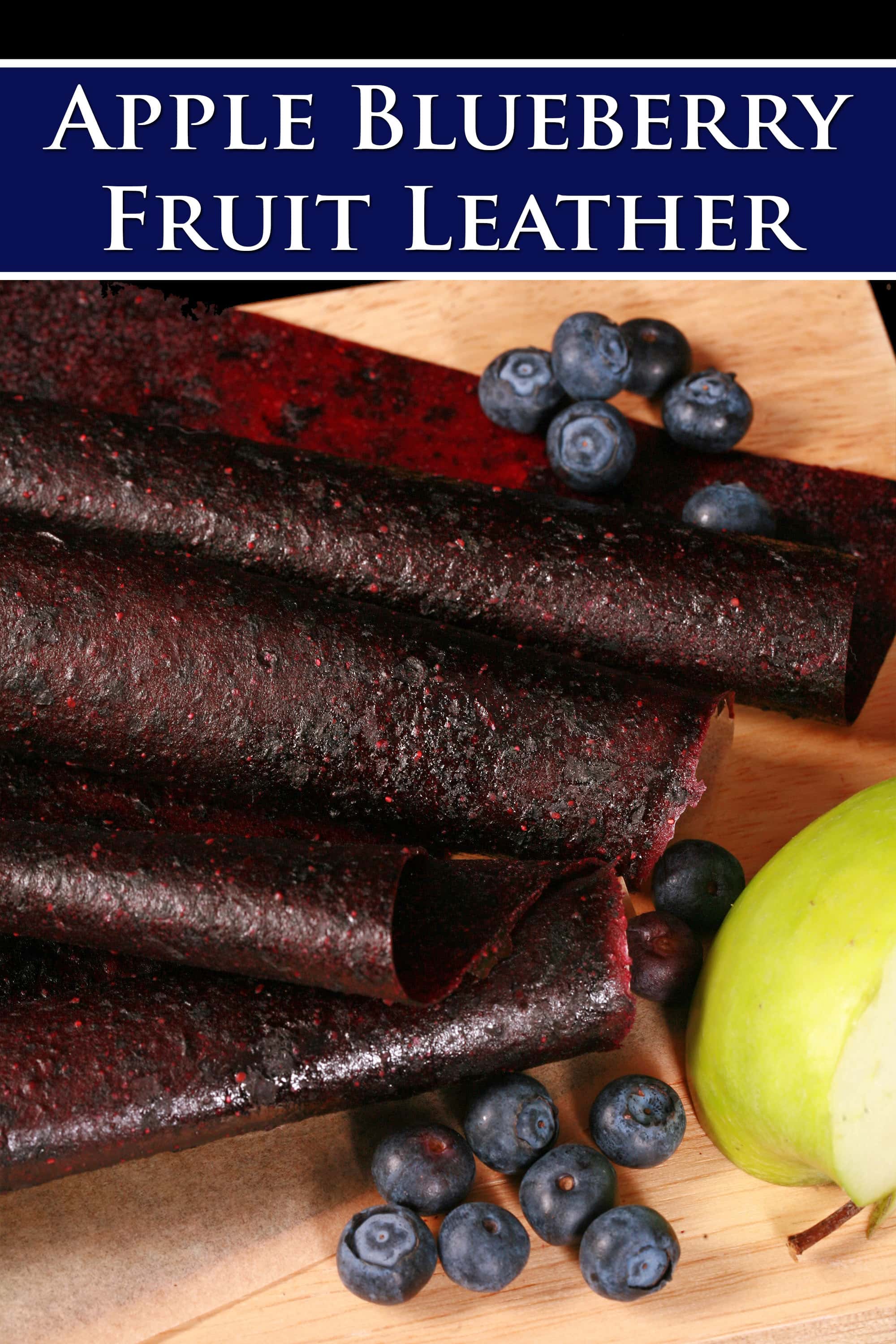 Several homemade blueberry apple fruit leather rolls on a cutting board, with a green apple and some fresh blueberries. Overlaid text says apple blueberry fruit leather.