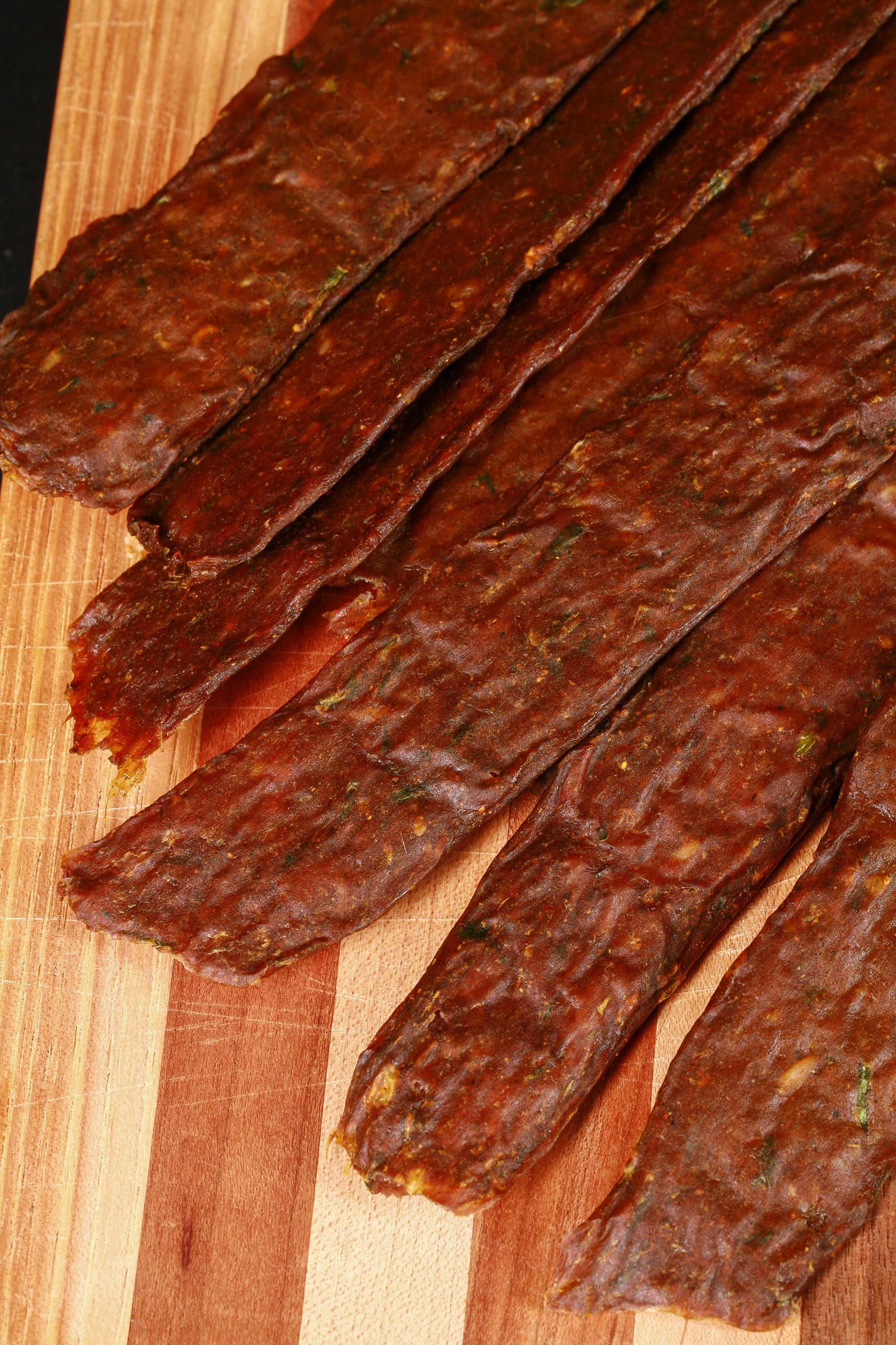 A pile of Trinidadian curry inspired chicken jerky sticks on a cutting board with green onions, garlic, and scotch bonnet peppers.