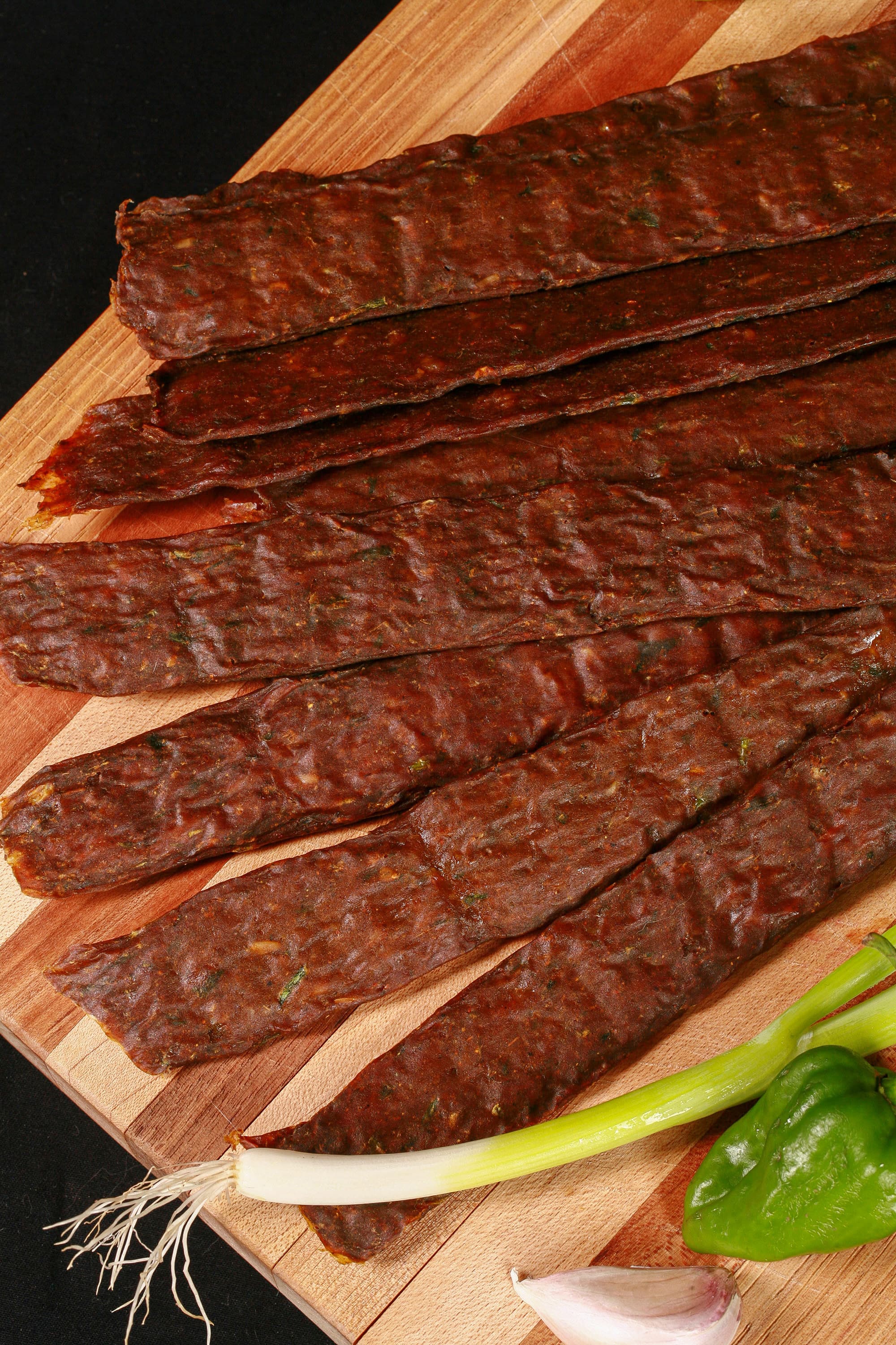 A pile of Caribbean curry chicken jerky sticks on a cutting board with green onions, garlic, and scotch bonnet peppers.