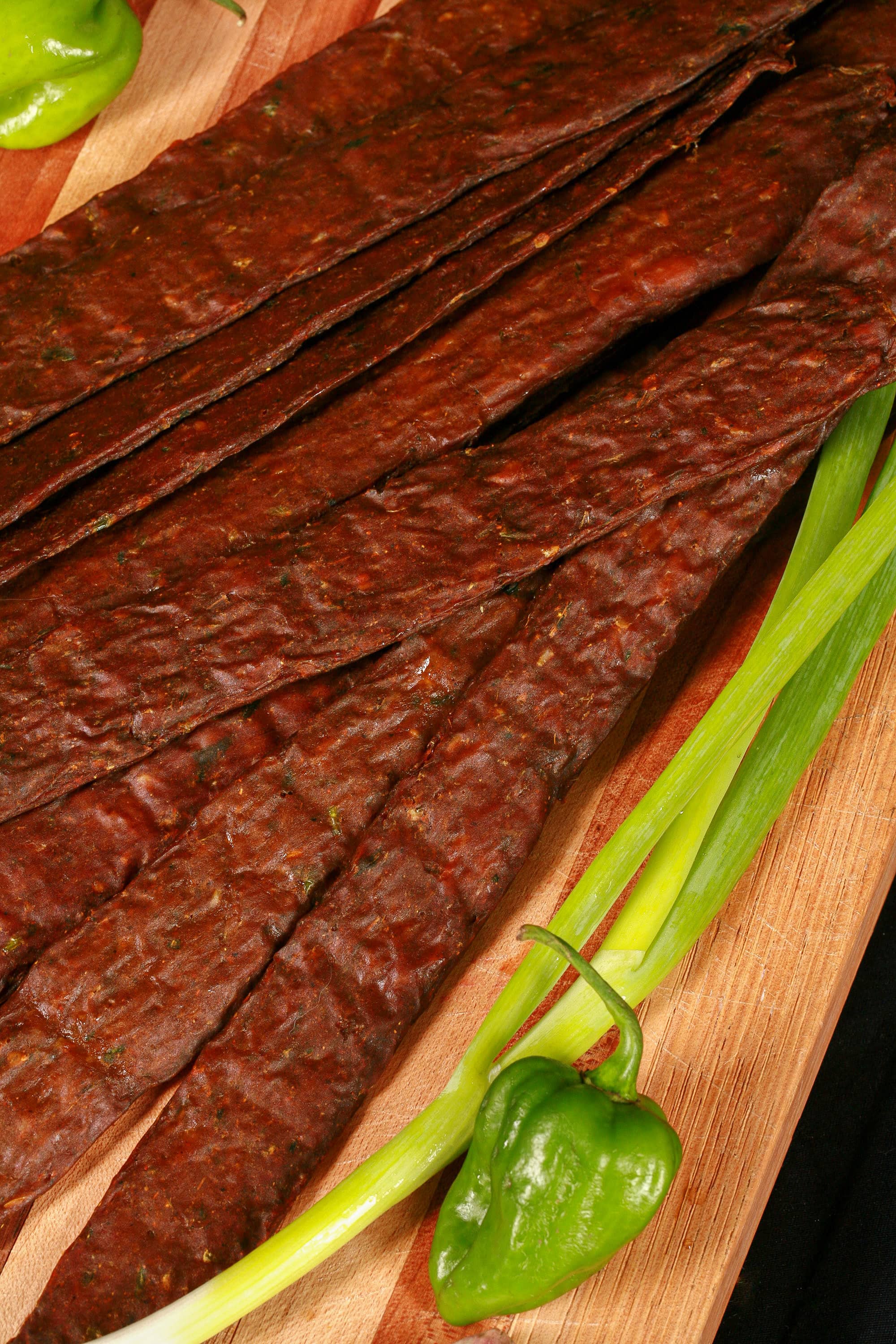 A pile of Caribbean curry chicken jerky sticks on a cutting board with green onions, garlic, and scotch bonnet peppers.