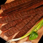 A pile of Caribbean curry chicken jerky sticks on a cutting board with green onions, garlic, and scotch bonnet peppers.