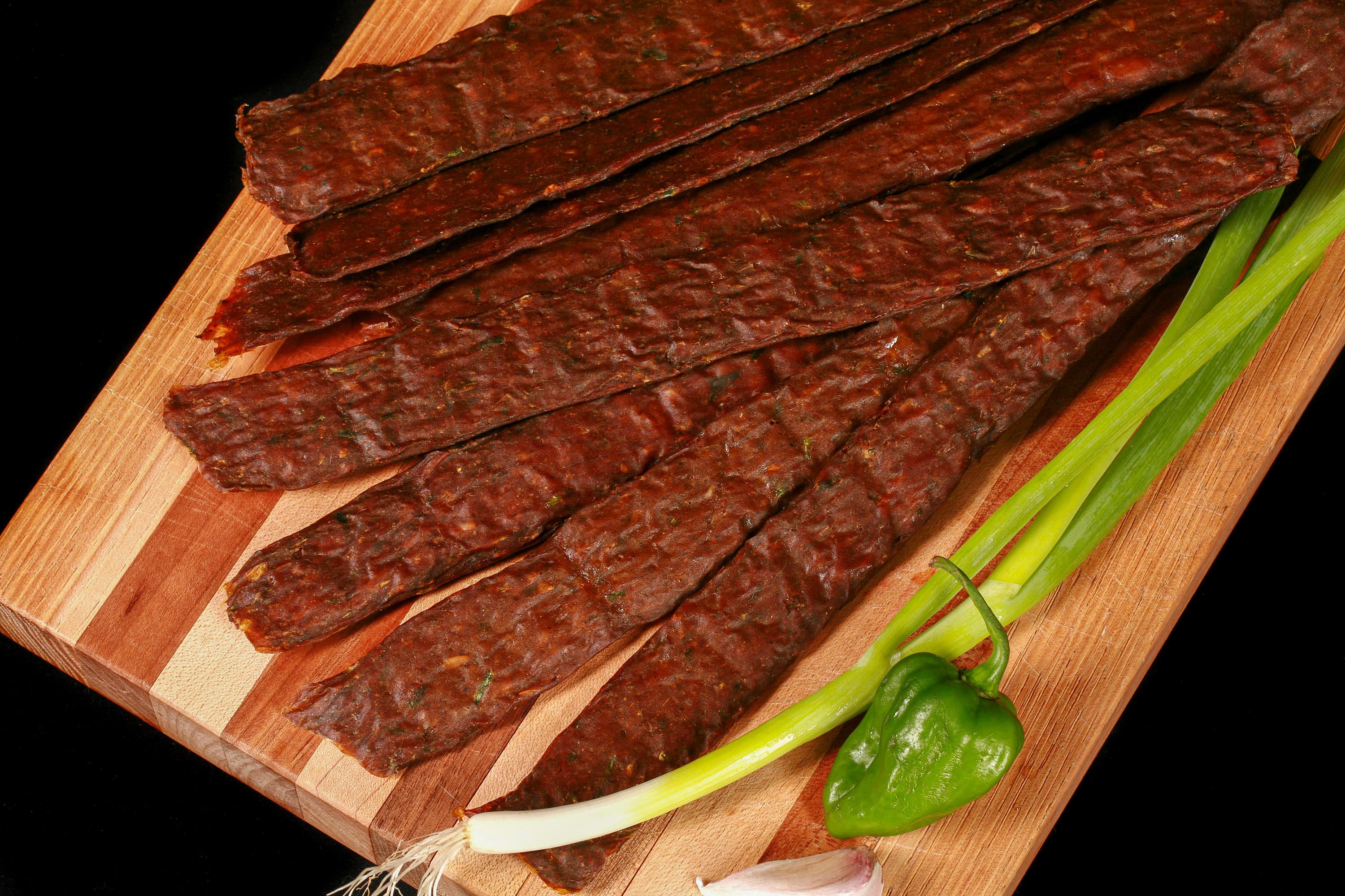A pile of Caribbean curry chicken jerky sticks on a cutting board with green onions, garlic, and scotch bonnet peppers.