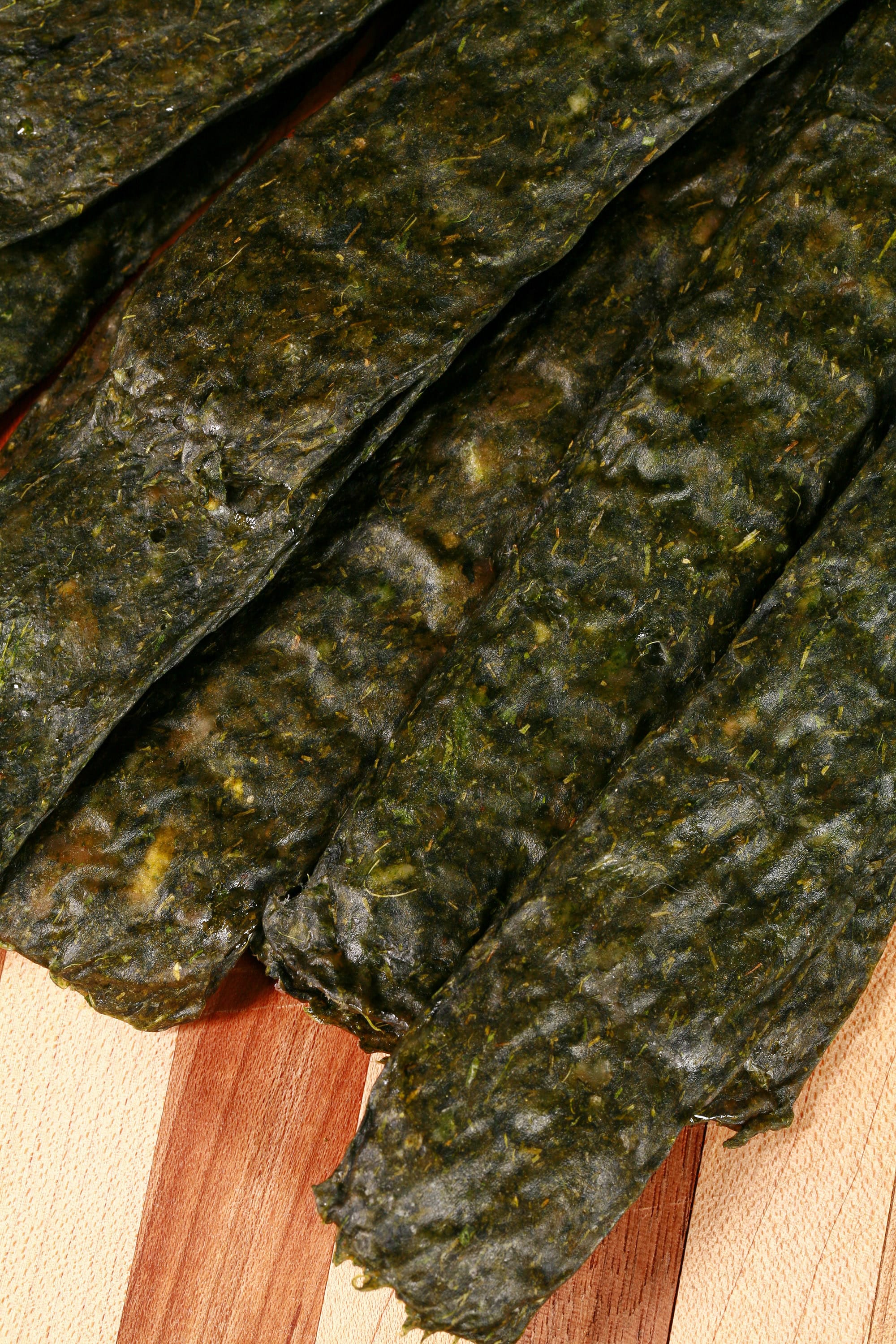 A pile of dark green spanakopita chicken jerky, with a lemon wedge, garlic, and fresh dill on a cutting board.
