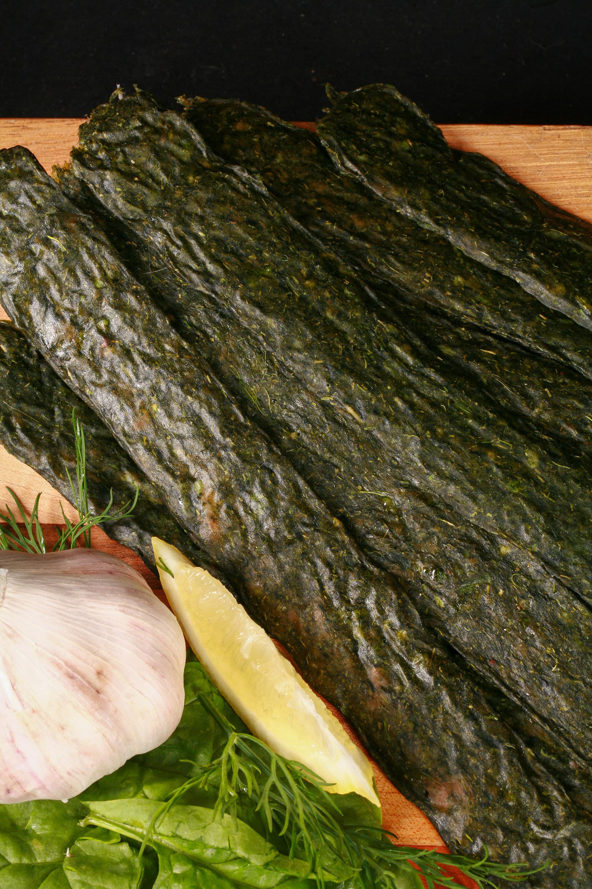 A pile of dark green spinach chicken jerky, with a lemon wedge, garlic, and fresh dill on a cutting board.