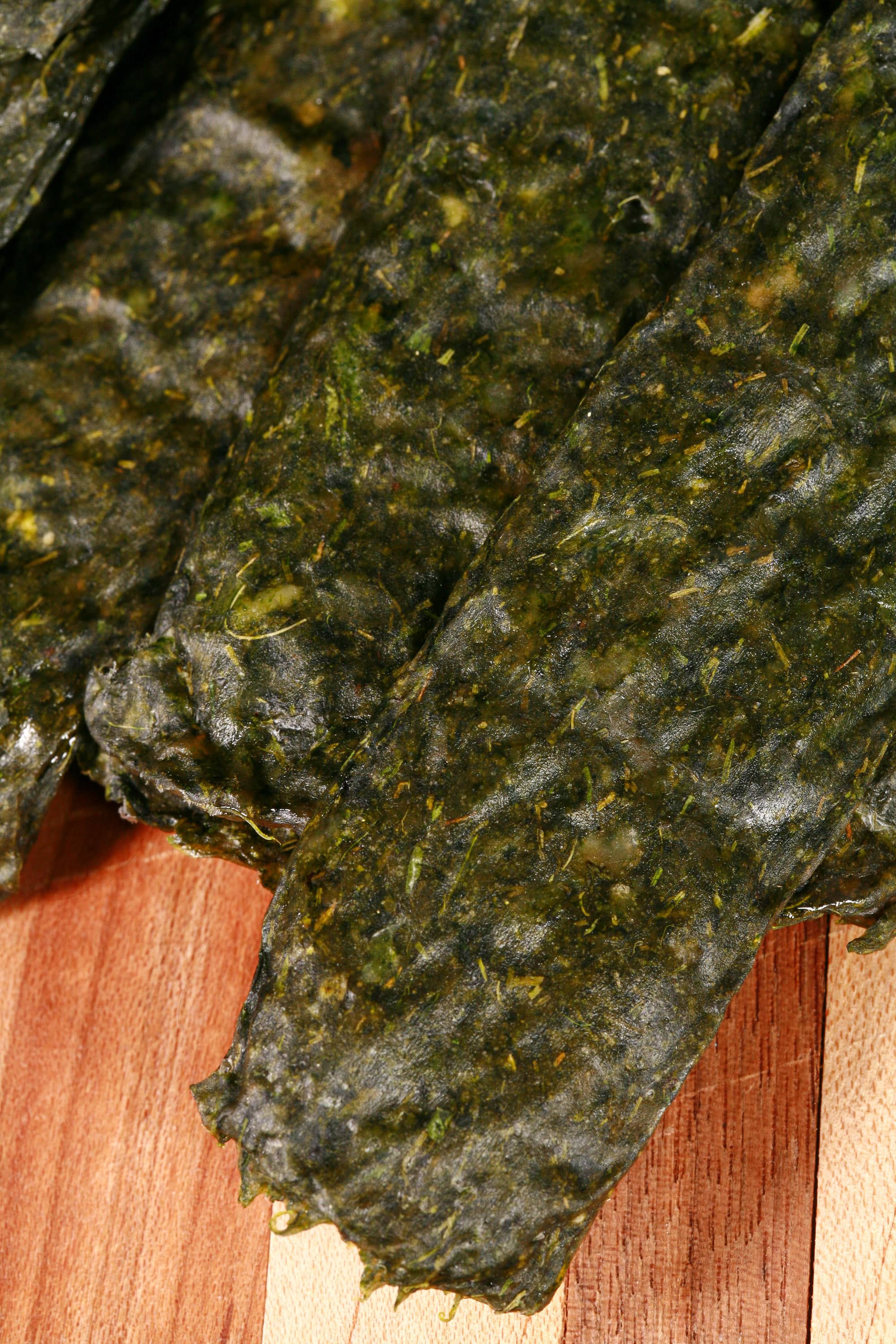 A pile of dark green spanakopita chicken jerky, with a lemon wedge, garlic, and fresh dill on a cutting board.