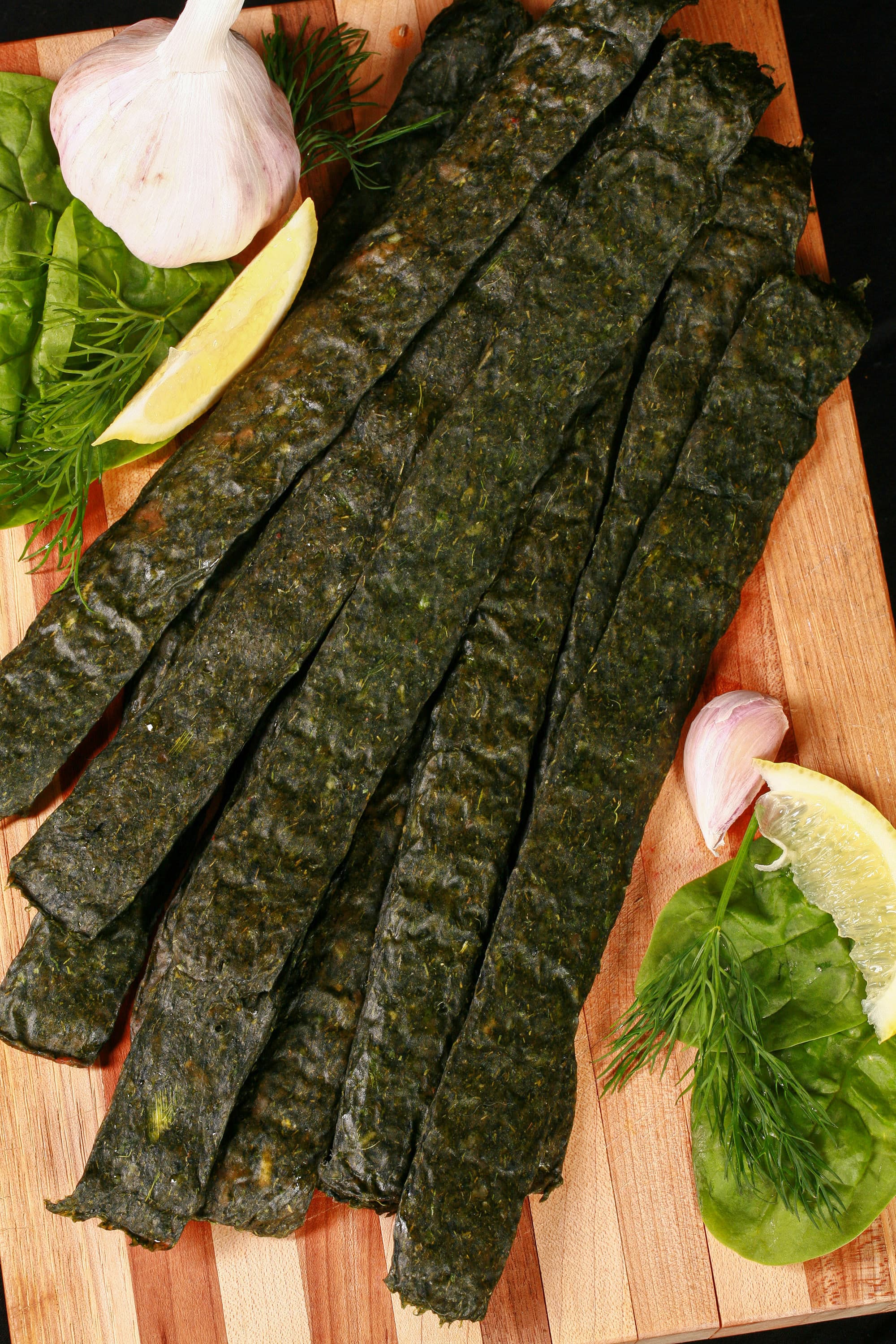 A pile of dark green spinach chicken jerky, with a lemon wedge, garlic, and fresh dill on a cutting board.
