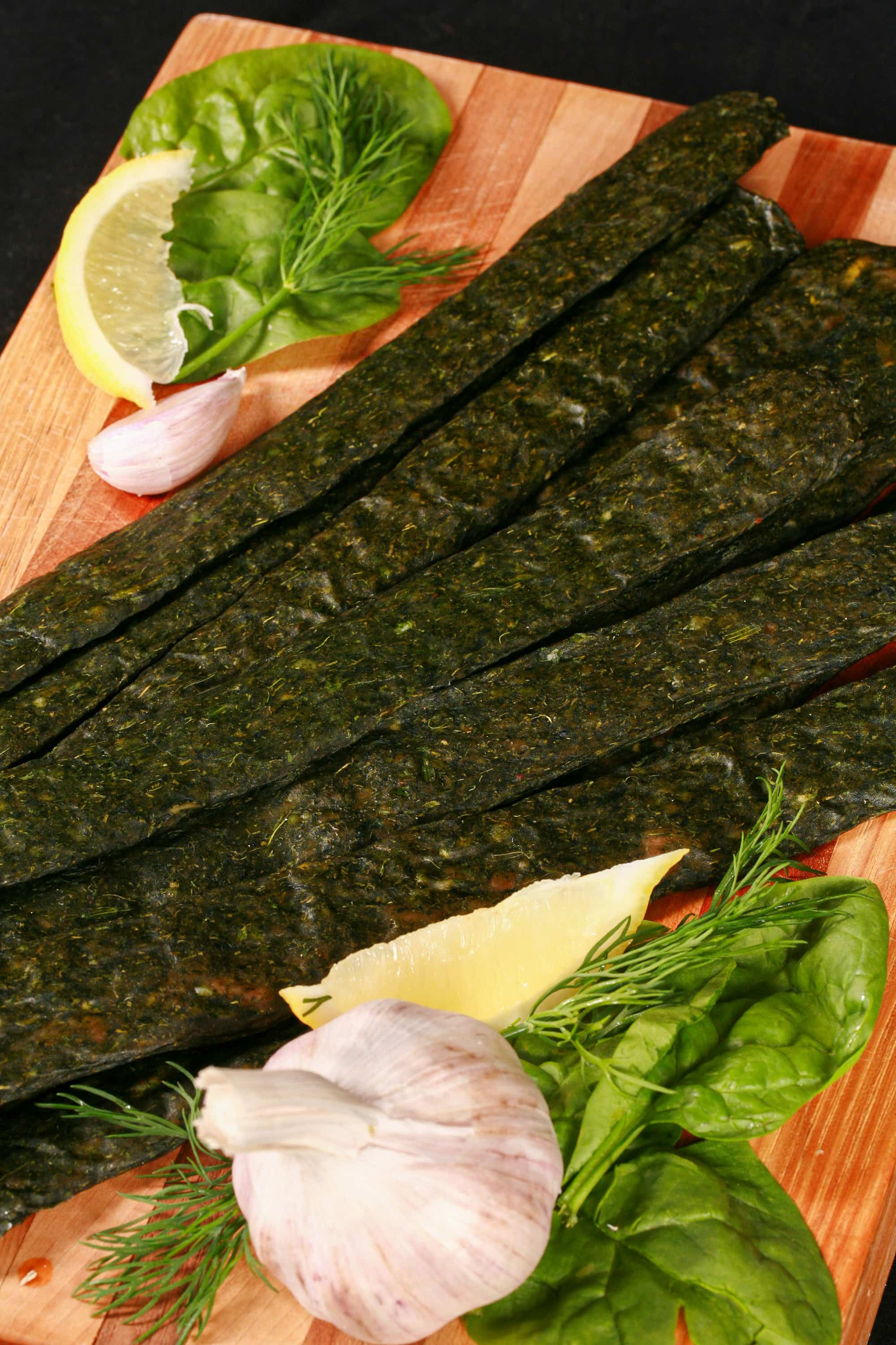 A pile of dark green spanakopita chicken jerky, with a lemon wedge, garlic, and fresh dill on a cutting board.