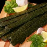 A pile of dark green spinach chicken jerky, with a lemon wedge, garlic, and fresh dill on a cutting board.
