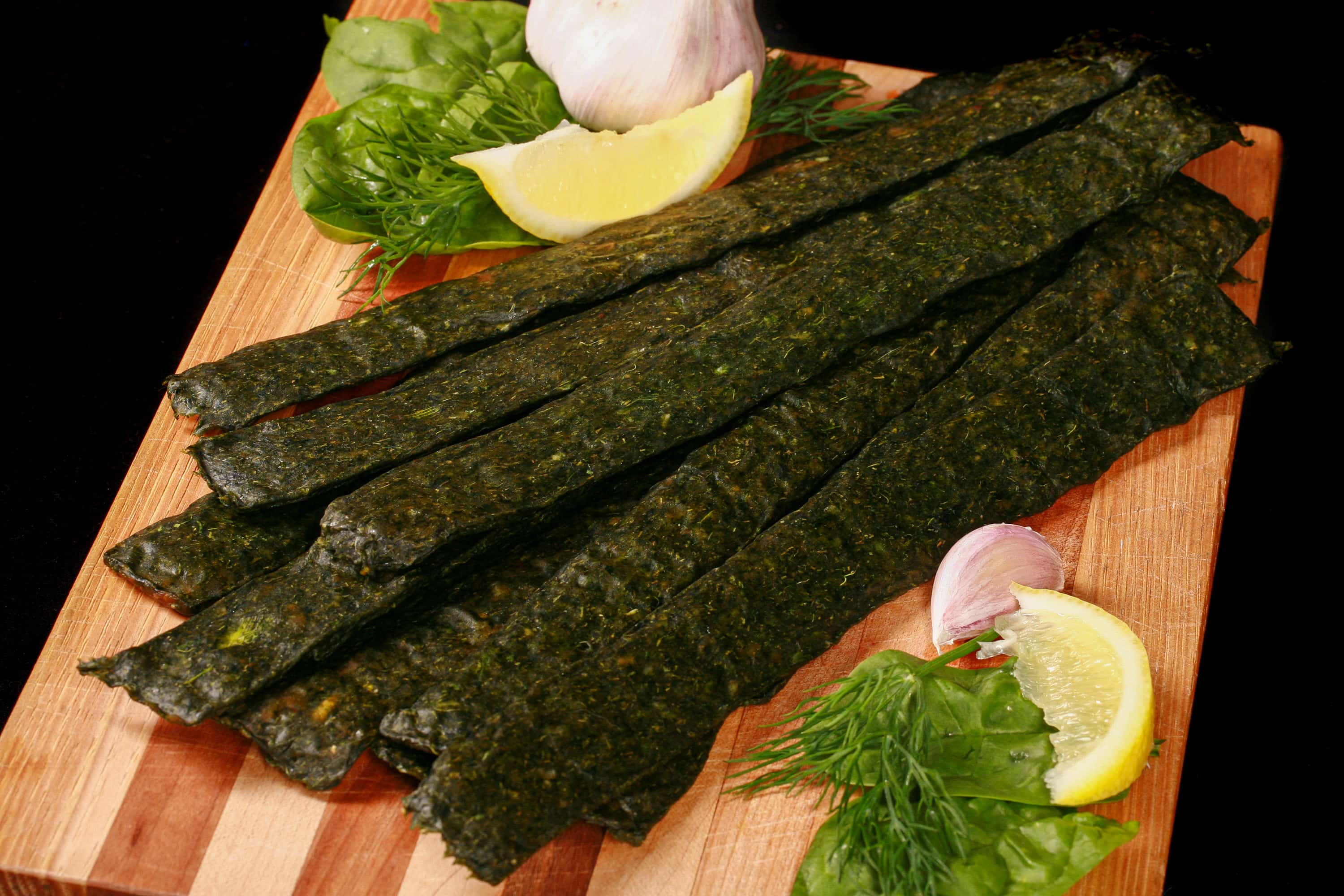 A pile of dark green spinach chicken jerky, with a lemon wedge, garlic, and fresh dill on a cutting board.