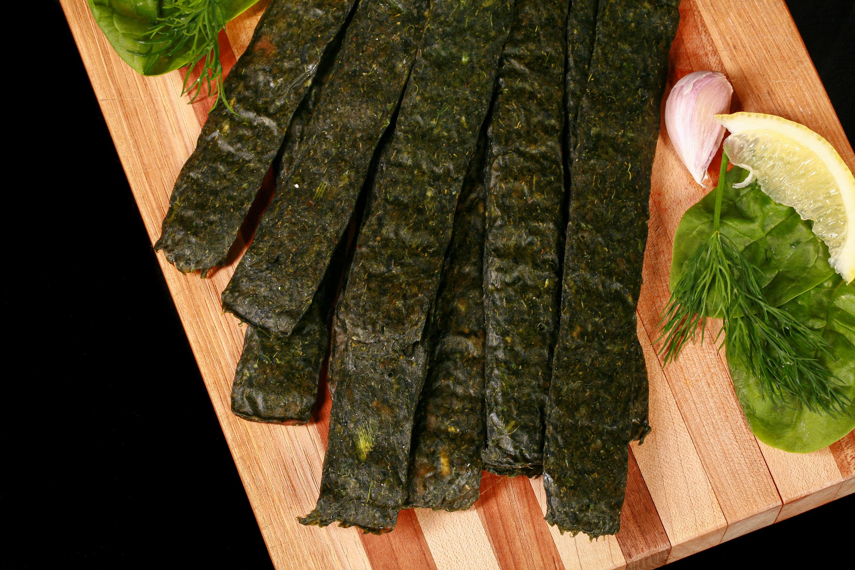 A pile of dark green spanakopita chicken jerky, with a lemon wedge, garlic, and fresh dill on a cutting board.